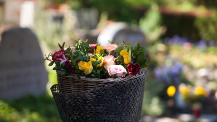 Korbtasche mit bunten Rosen steht auf einem Grab, im Hintergrund sind unscharfe Blumen und Grabsteine sichtbar.