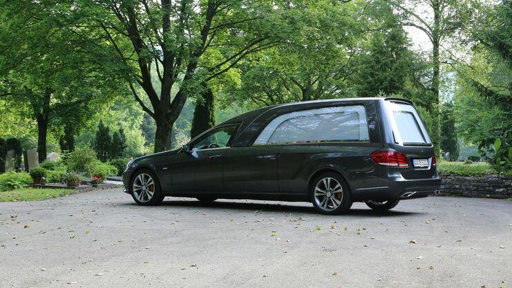Dunkler Leichenwagen parkt auf einem Weg zwischen Bäumen auf einem Friedhof, neben einer Bank.