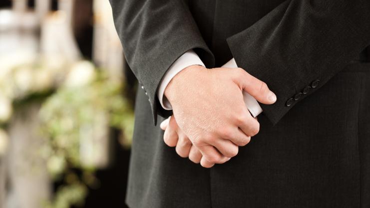 Ein Mann in einem Anzug steht mit verschränkten Händen vor einer dekorierten Kulisse mit Blumen und Kerzen.