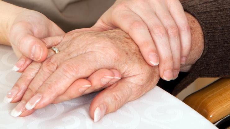 Zwei Hände, eine ältere und eine jüngere, liegen auf einem Tisch, wobei die jüngere Hand die ältere sanft umschließt.