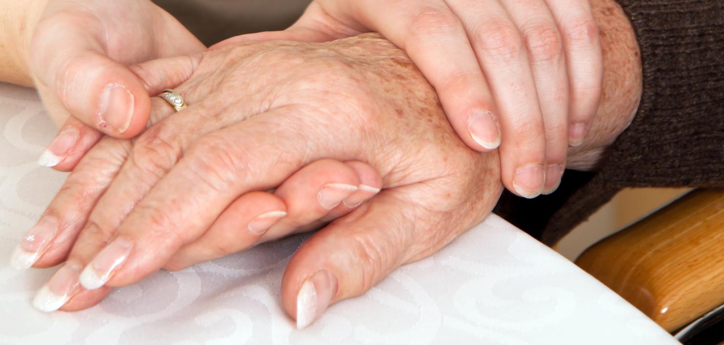 Zwei Hände, eine ältere und eine jüngere, liegen auf einem Tisch, wobei die jüngere Hand die ältere sanft umschließt.